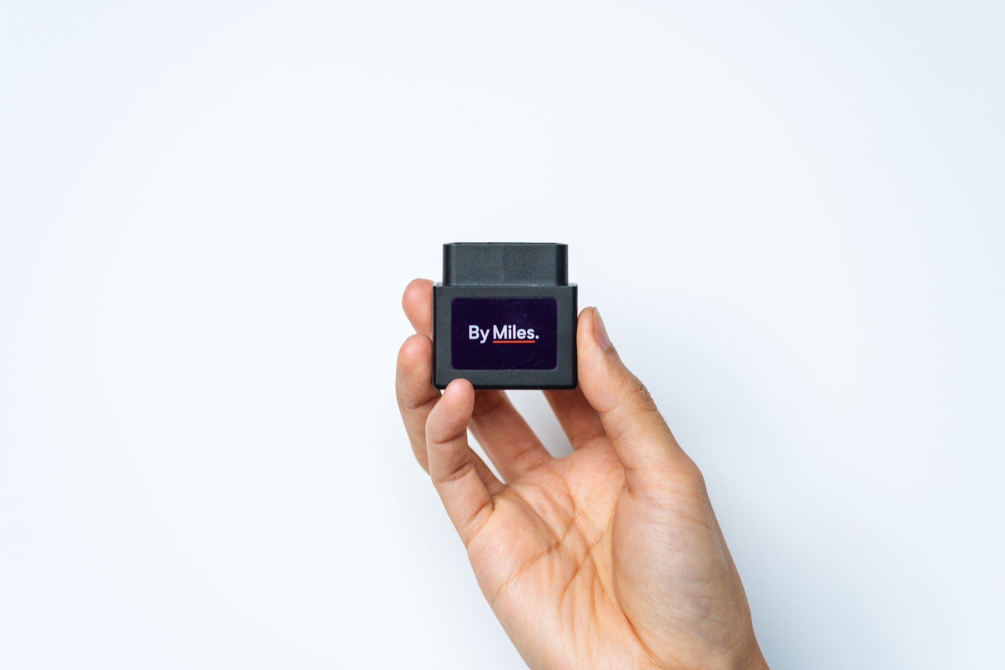Hand holding a By Miles Tracker device against a white background. The small black device measures miles driven for pay-per-mile car insurance.
