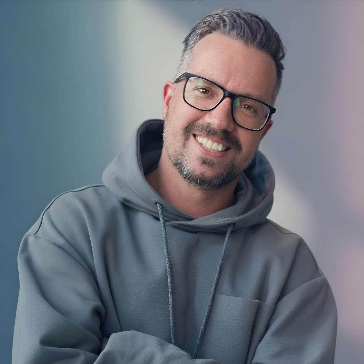 Man with black-rimmed glasses and gray hoodie, smiling warmly against a professional gray background. Salt-and-pepper hair and beard.